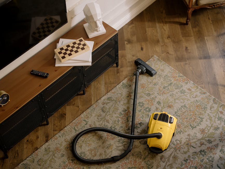 A yellow cylinder vacuum cleaner with a black hose attachment is positioned on a patterned area rug, cleaning a wooden floor in a living room. The room features a black metal and wood sideboard against the wall, topped with a white bust sculpture, a remote control, and a stack of papers with a small chessboard. The lighting is natural, casting soft shadows, and the surfaces appear clean and well-maintained, highlighting professional surface cleaning and deep cleaning techniques used by Ruislip Carpet Cleaning.