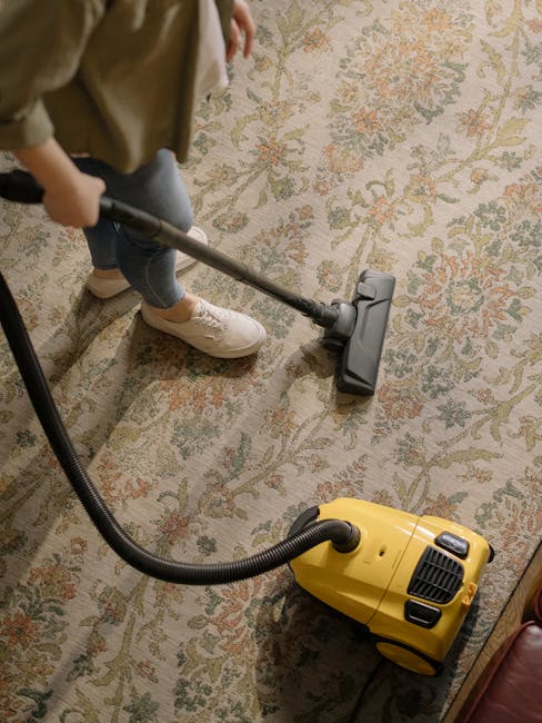 A person using a vacuum cleaner on a patterned, multi-colored carpet in a residential room. The vacuum cleaner is yellow with a black hose, and the individual is wearing beige shoes, blue jeans, and a beige jacket. The carpet features a traditional floral design with cream, green, orange, and pink hues. The scene is well-lit, emphasizing the clean and maintained condition of the carpet. This setup illustrates practical surface cleaning as part of domestic cleaning services offered by Ruislip Carpet Cleaning. The image highlights the use of professional equipment to achieve thorough deep cleaning and sanitisation of residential carpets.
