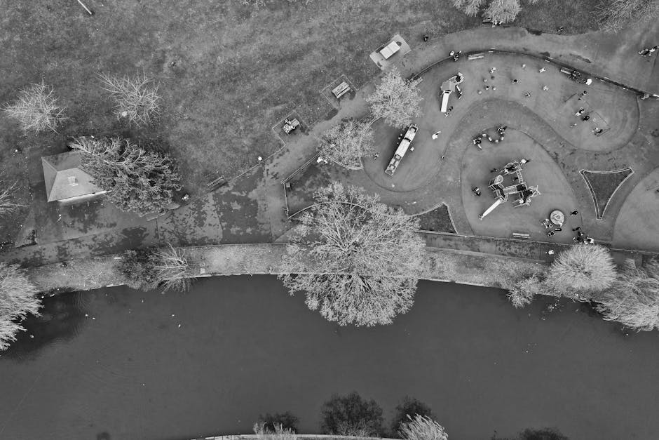An aerial black-and-white view of a park surrounding a body of water, showing a playground with curved pathways and various play equipment, trees with bare branches indicating winter, and a small shelter or pavilion near the water's edge. The scene is clean and orderly, with no visible debris or dirt, reflecting high standards of surface cleaning and maintenance typical of professional cleaning services like those offered by Ruislip Carpet Cleaning for domestic and commercial environments.