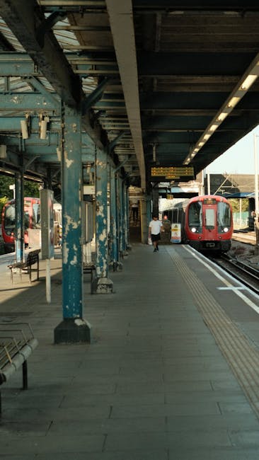 Exterior view of South Ruislip railway station platform during daytime, featuring weathered blue-painted steel supports, a concrete tiled walkway, and a red train arriving or departing on the tracks. A single passenger walks along the platform, which is illuminated by natural light and equipped with benches and safety tactile paving. The station is clean and maintained, providing a basic shelter for travelers. Ruislip Carpet Cleaning emphasizes surface cleaning and maintenance, similar to the sanitation processes required for residential and commercial spaces, ensuring hygiene and cleanliness. The image reflects the station's functional design, with minimal clutter and clear pathways, aligning with professional cleaning and upkeep standards.