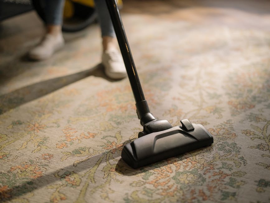 Close-up image of a vacuum cleaner head actively cleaning a patterned area rug with intricate floral designs in shades of beige, green, and orange. The vacuum head is black with a grey button, attached to a black wand, and is positioned centrally on the carpeted floor. In the background, legs of a person in dark trousers and white shoes are visible, standing on the rug, suggesting active surface cleaning. The lighting is natural, highlighting the clean and well-maintained appearance of the woven textile. This scene exemplifies domestic surface cleaning performed by Ruislip Carpet Cleaning, emphasizing their expertise in deep carpet and rug cleaning services near South Ruislip station.